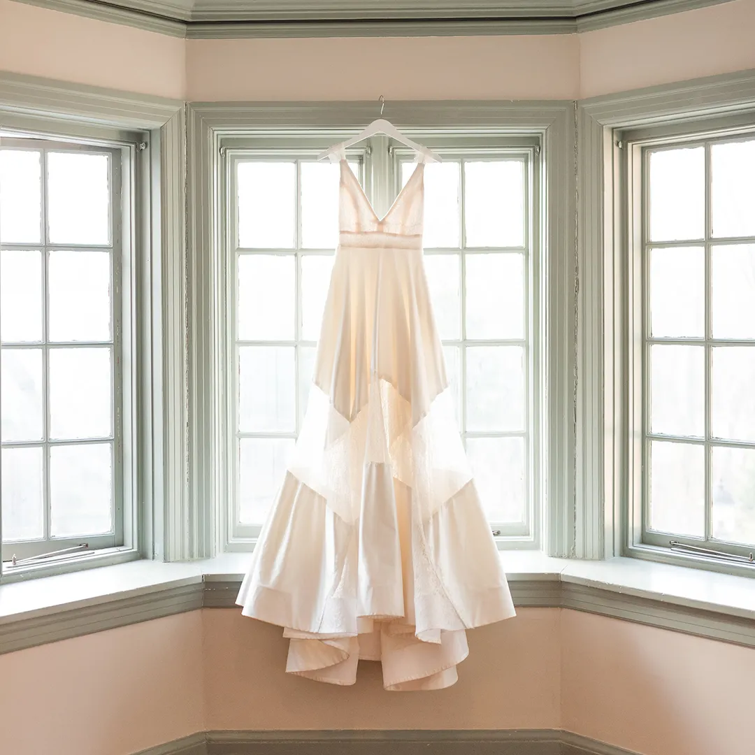 Elegant white wedding dress with lace details hanging on a hanger in a corner window with light gray trim.
