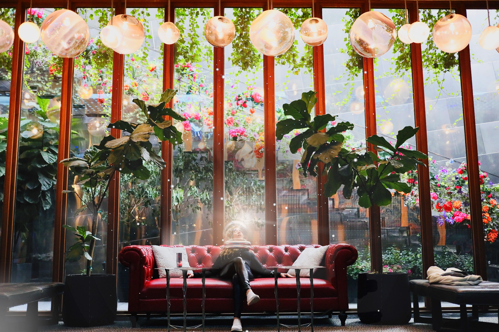 Person wearing an astronaut helmet sitting on a red leather sofa in a bright room with large windows, hanging plants, and round ceiling lights.
