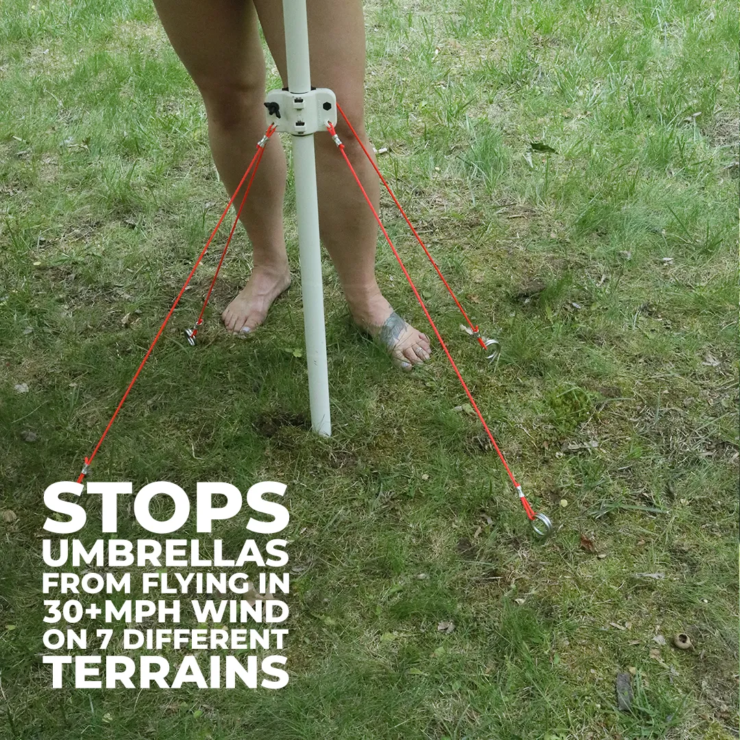 Bare feet standing on grass securing an umbrella pole with red tension cords attached to ground anchors.