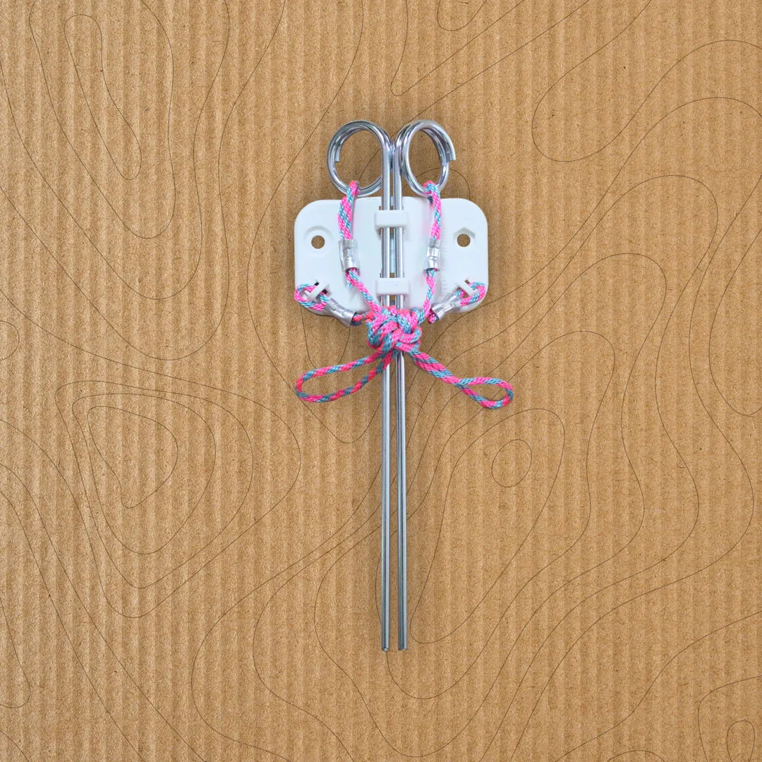 Metal anchor with two prongs and pink and blue cord tied in a bow, mounted on a white plastic base against a corrugated cardboard background.